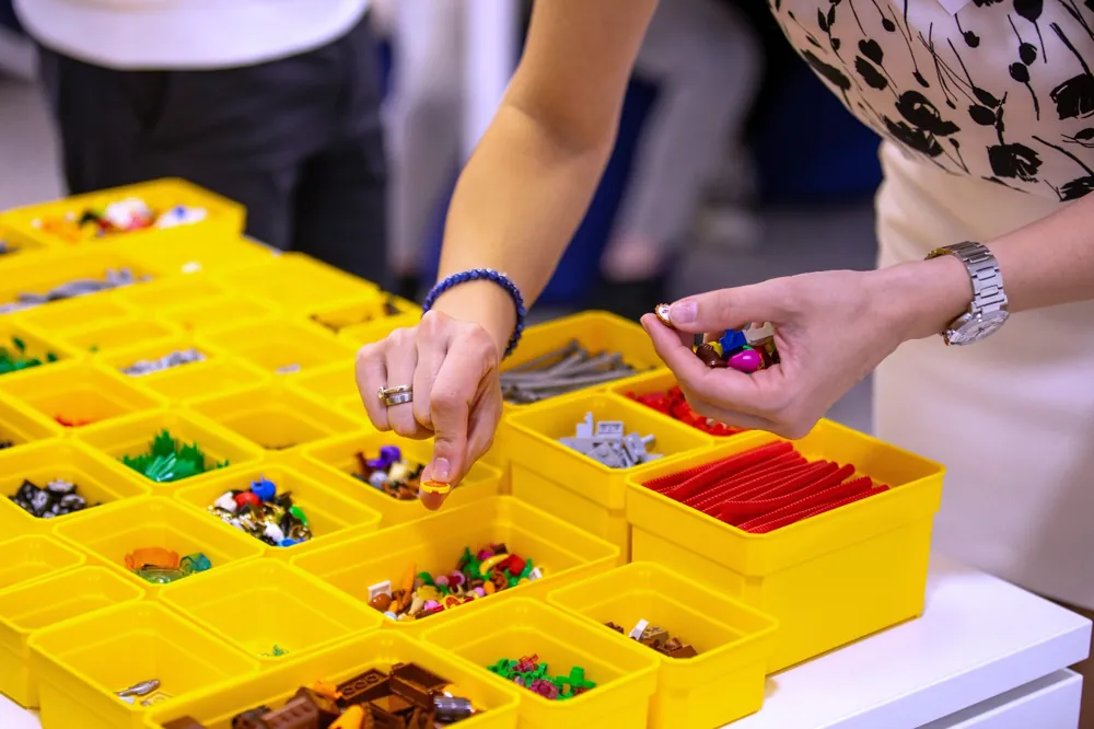 Două perechi de mâini aleg piese dintr-un set de cutii galbene cu elemente LEGO sortate, la începutul unui workshop.