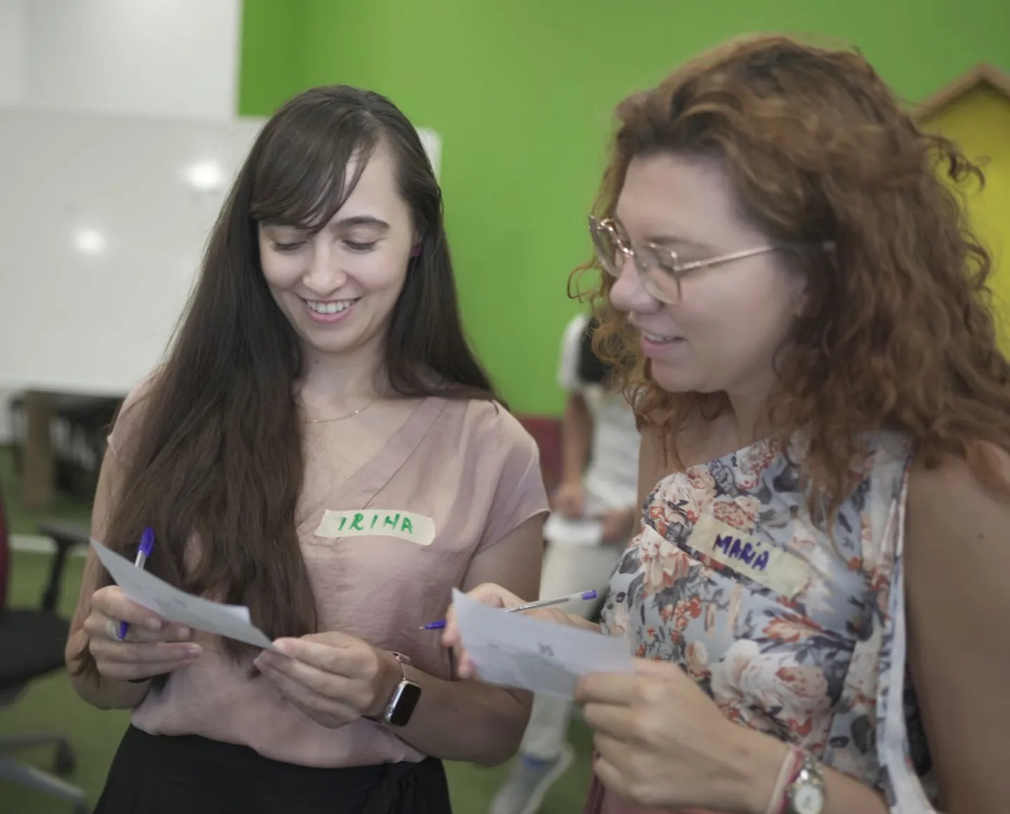 Două participante la un workshop, cu ecusoane scrise de mână, fiecare cu un bilet de hârtie și un pix; una notează un punctaj pe biletul primit.